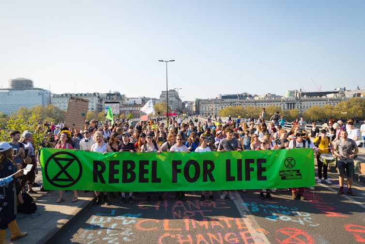 Protesters march across the bridge holding a large green sign with the Rebellion Extinction logo and the words 