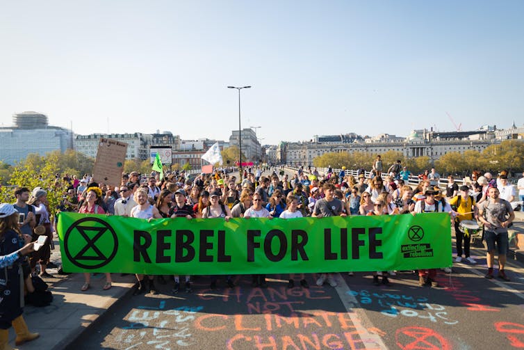 Protesters march across the bridge holding a large green sign with the Rebellion Extinction logo and the words