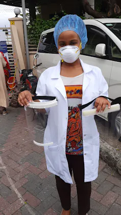 A woman wearing a lab coat, face mask and hair covering holds two plastic face shields
