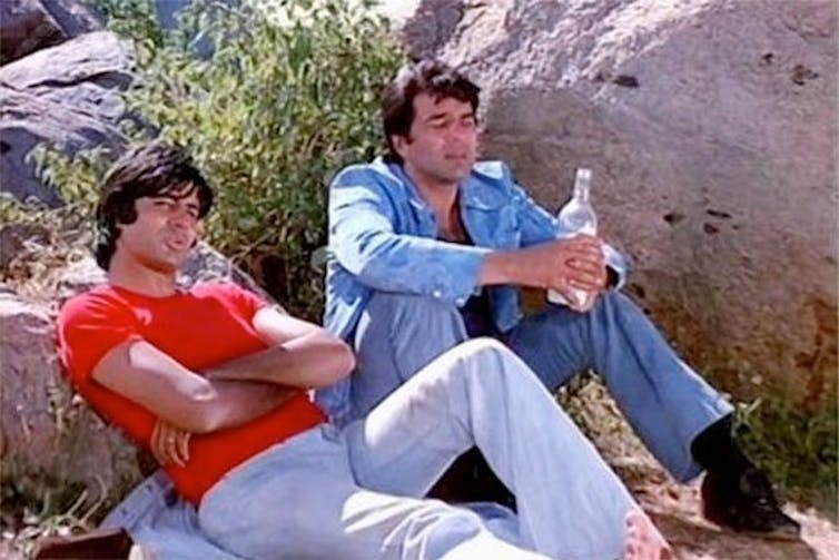 Two men lean against a rock. Both wear jeans.