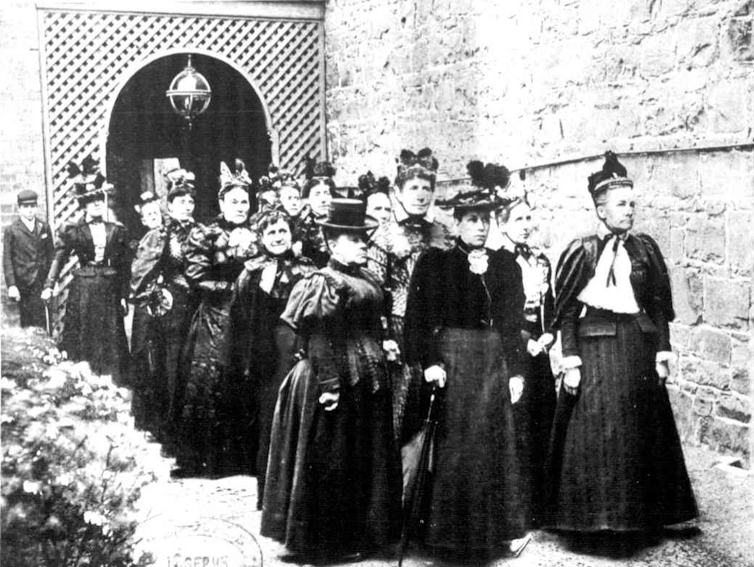 13 women in rows of three, in stiff formal wear of the time.