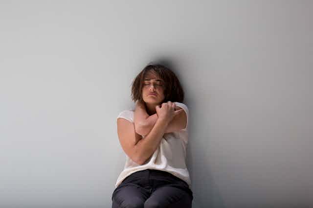 A woman leans against a wall, hugging herself.