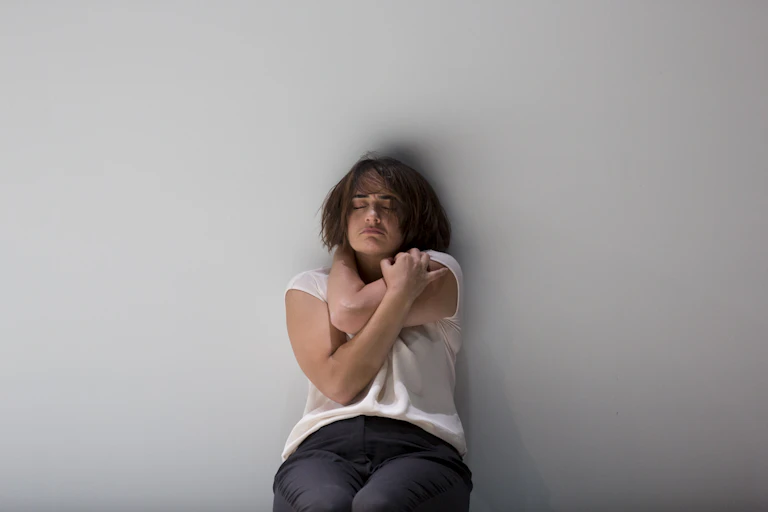 A woman leans against a wall, hugging herself.
