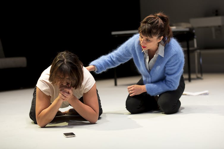 A woman kneels on the ground and cries, comforted by a friend.