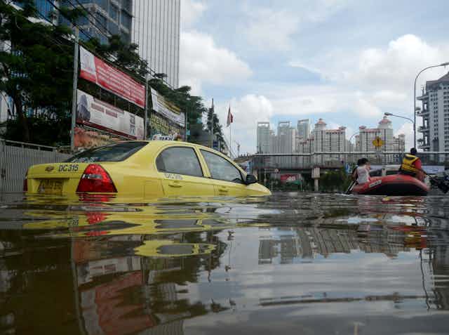 Taksi berwarna kuning tenggelam oleh banjir dengan gedung sebagai latar belakang. 