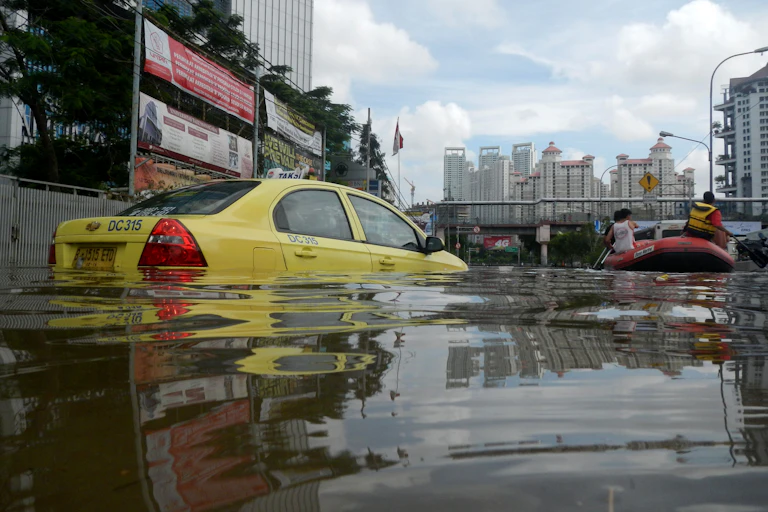 Taksi berwarna kuning tenggelam oleh banjir dengan gedung sebagai latar belakang.