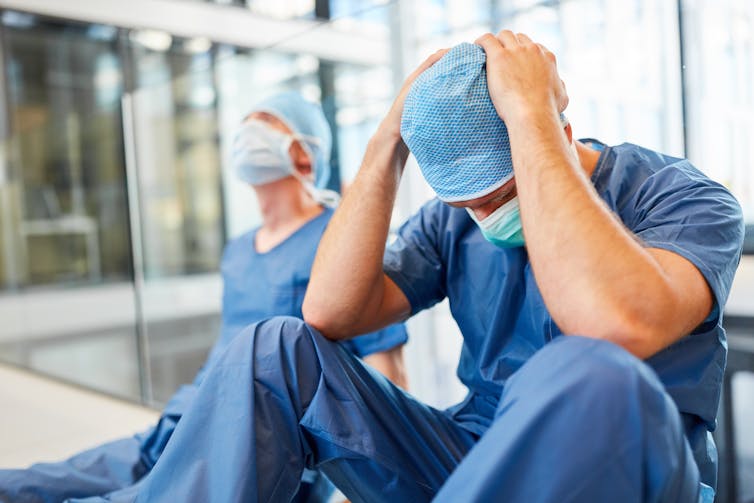 Dos sanitarios sentados en el suelo  de un pasillo de hospital con gesto de cansancio.