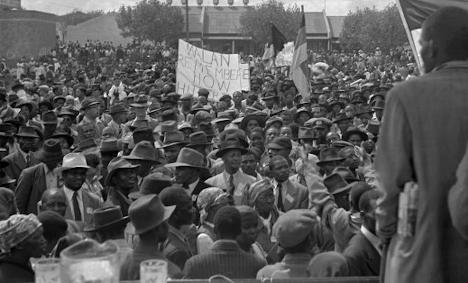 A man in a suit, his back to the camera, addresses a large crowd.