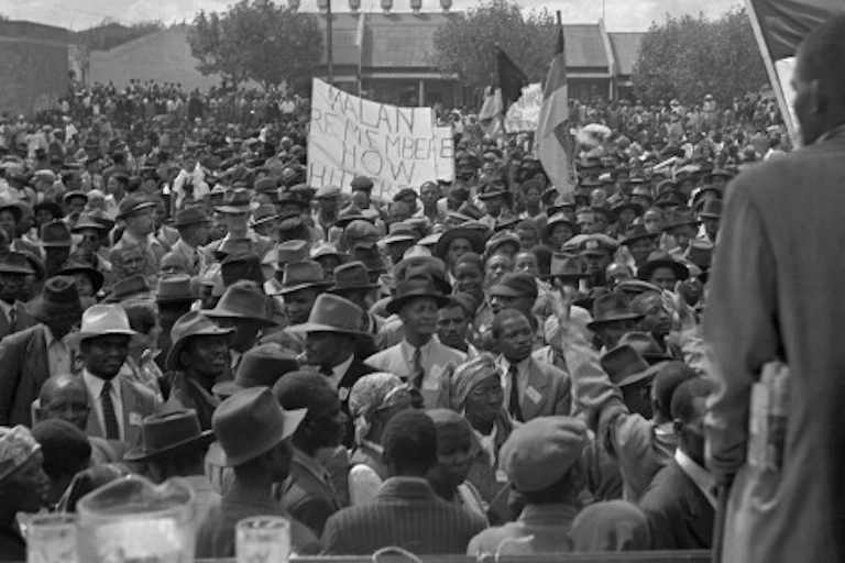 A man in a suit, his back to the camera, addresses a large crowd.