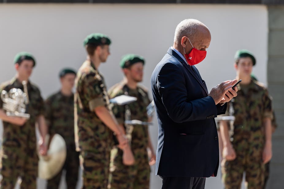 Swiss Federal councillor Ueli Maurer looks at his phone while members of the military wait in the background.