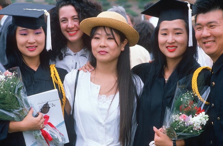 Two Asian American women in graduation gowns pose with other people