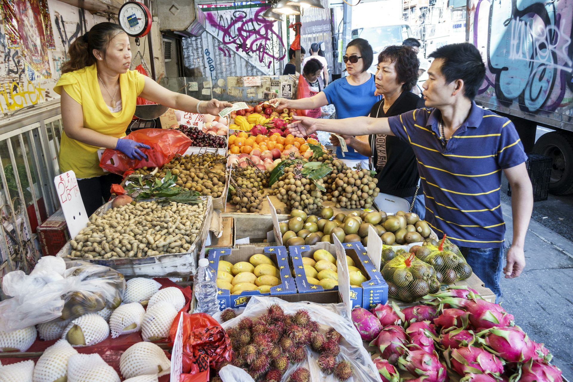 Asian woman hands change back to three Asian American customers, over a large outdoor fruit display