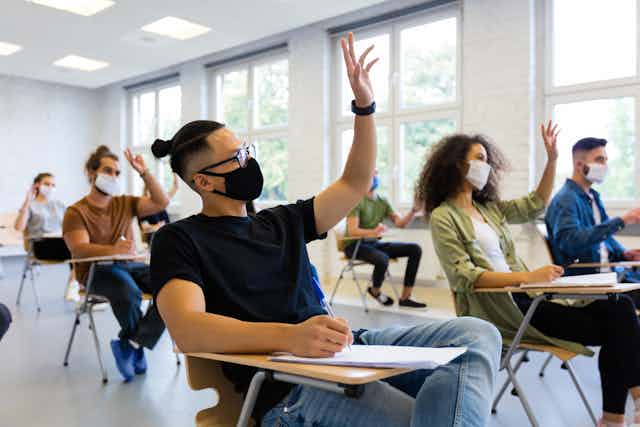 A group of masked college students raise their hands in class.