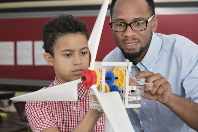 An African American students and his science teacher use a model wind turbine together in class.