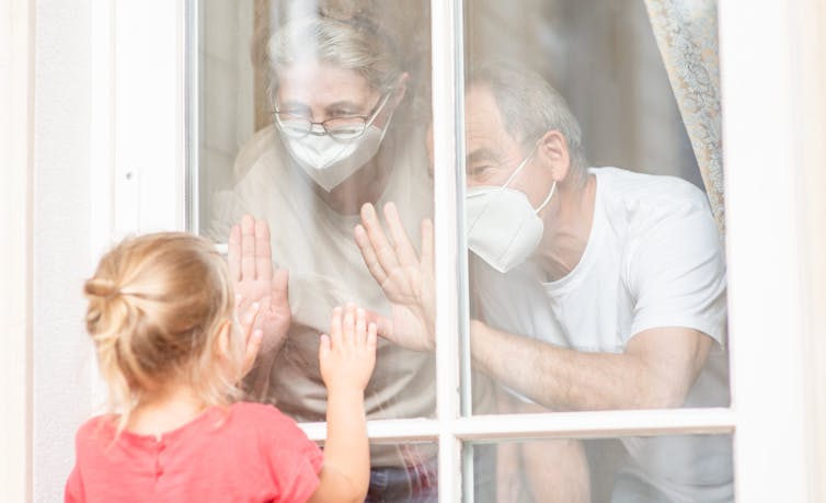 Grandparents with masks seen pressing hands against window looking at granddaughter