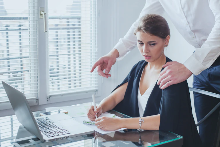 A woman sits as a desk with an uncomfortable look as a man standing behind puts his left hand on her left shoulder
