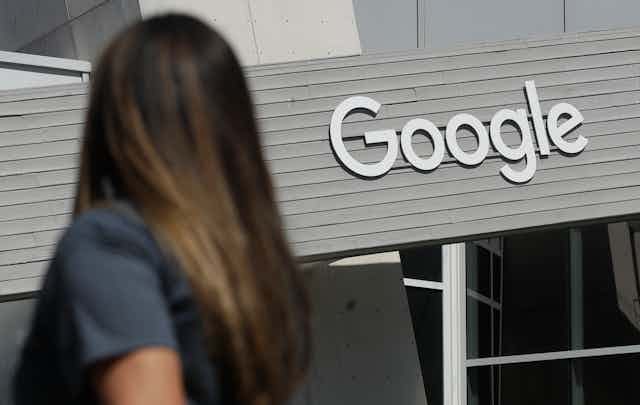 A woman is seen from behind as she walks past a building with a large Google sign.