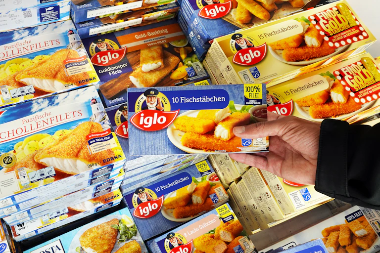 A hand holding a pack of fish fingers in a supermarket freezer aisle.