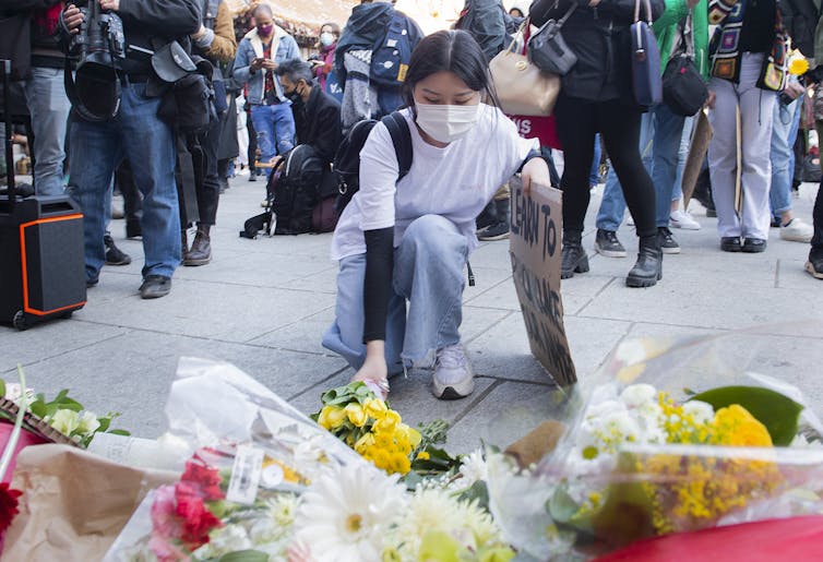 A woman kneels down to place flowers at a memorial. Behind her, a protest.