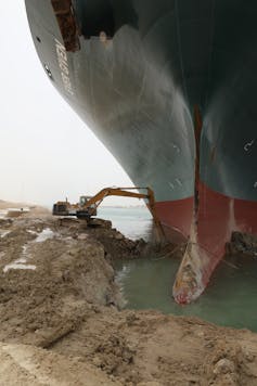 A digger extracting soil at the base of a large ship's hull