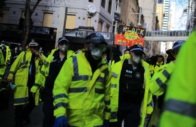 A group of police people in hi vis gear, walking in the street. There is a Black Lives Matter protest sign in the background.