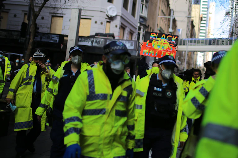 A group of police people in hi vis gear, walking in the street. There is a Black Lives Matter protest sign in the background.