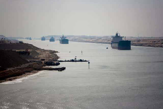 Cargo ships in single file moving along a narrow body of water between sandy banks.
