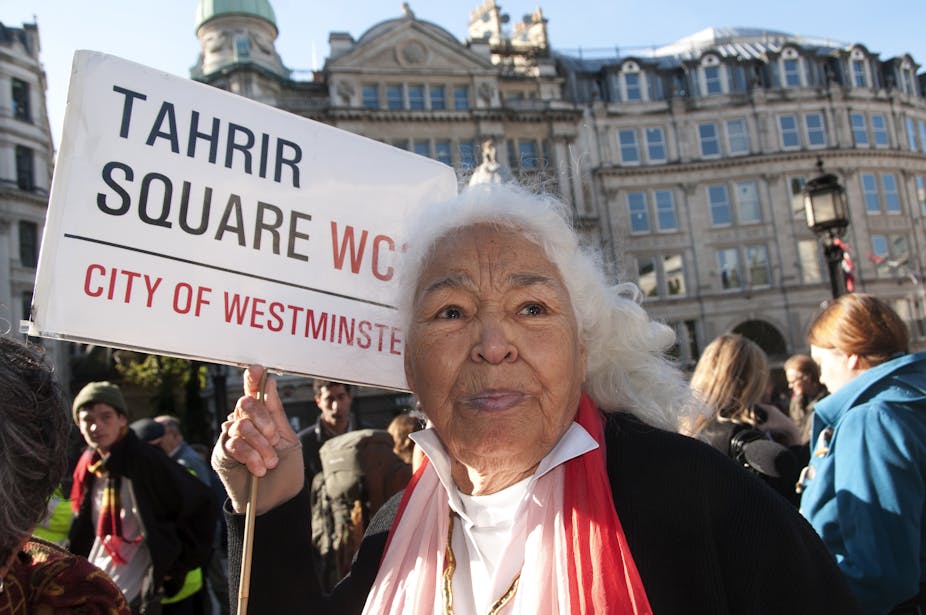 An elderly woman with white hair is in a crowd of people in front of grand European buildings. She holds a placard containing the words 'Tahrir Square'.