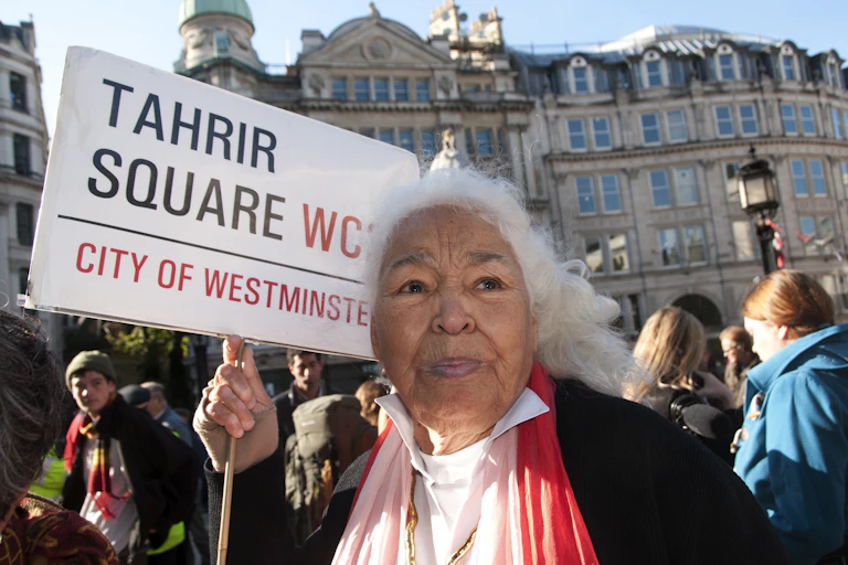 An elderly woman with white hair is in a crowd of people in front of grand European buildings. She holds a placard containing the words 'Tahrir Square'.