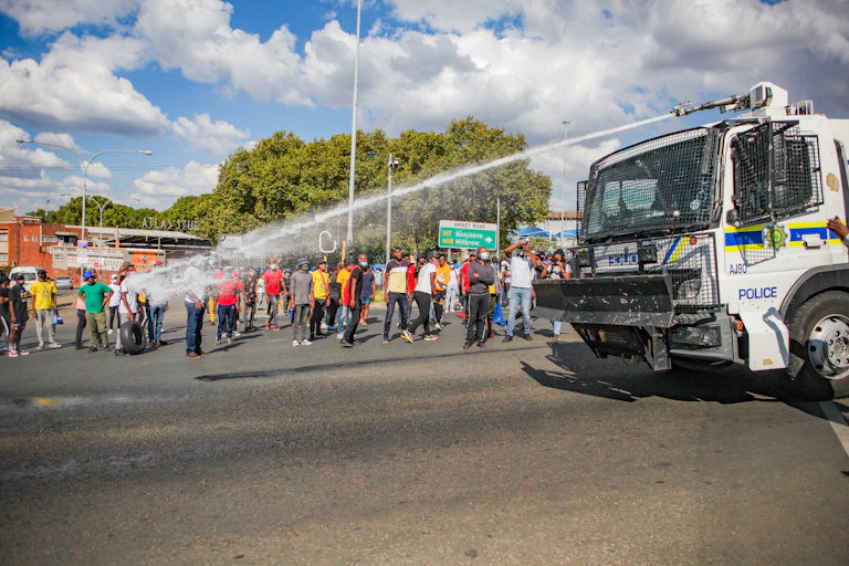 Protesting students standing on the sidewalk looking on at a police truck with a water cannon on the road.