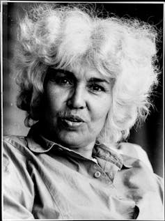 A woman looks directly to camera, her white hair done in dramatic fashion, her lips appear to be talking.