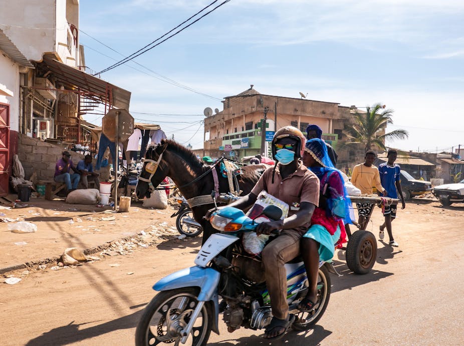 Une scène de rue à Mbour, au Sénégal, avec un homme et une femme sur un scooter et une charrette tirée par un cheval. 