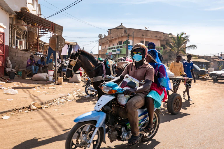 Une scène de rue à Mbour, au Sénégal, avec un homme et une femme sur un scooter et une charrette tirée par un cheval.