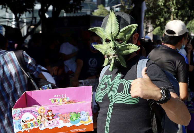 Man holding box of Kinder candies wears a marijuana leaf-shaped mask and gives a thumbs-up