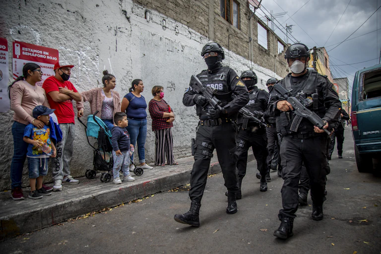Heavily armed soldiers in full riot gear pass a small crowd of women and children