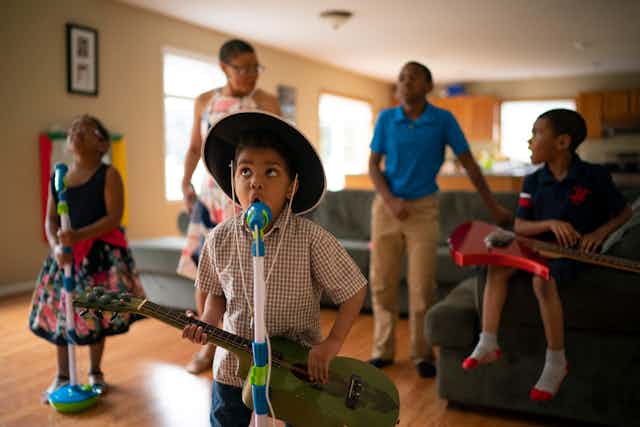 A young Black boy wearing a cowboy hat and holding a guitar sings into a microphone in his living room.