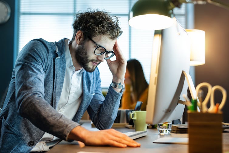 A young man sits at his computer. He looks stressed.