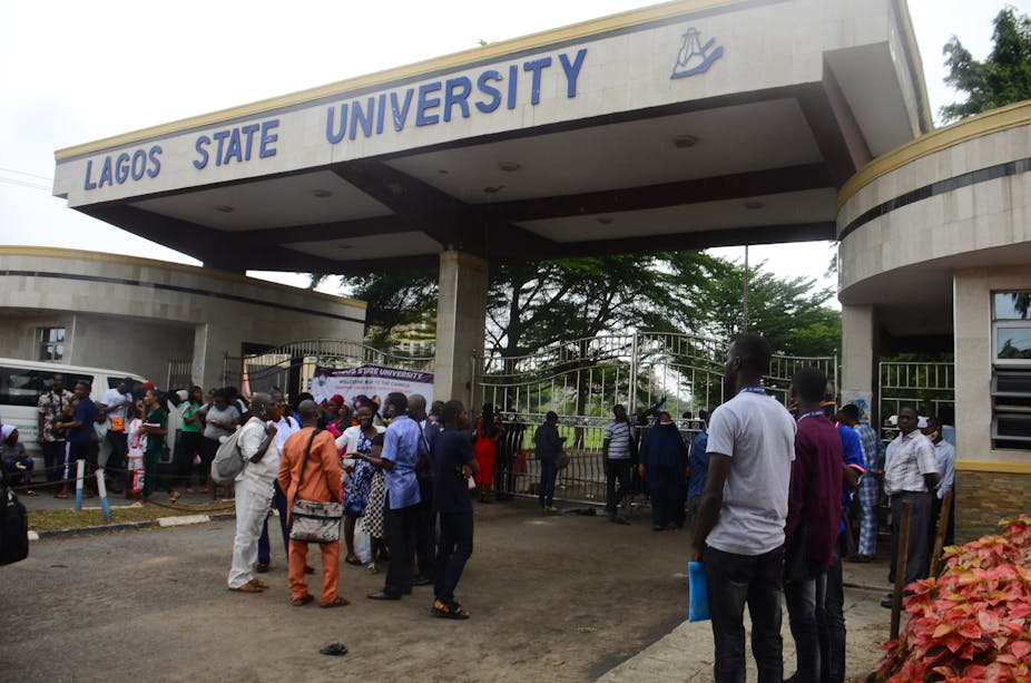 People stand outside a closed gate under a roof structure with a sign that reads Lagos State University
