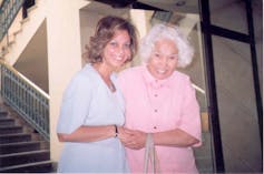 Dressed in pale blue, a smiling young woman holds the arm of a grey-haired older woman dressed in pink.