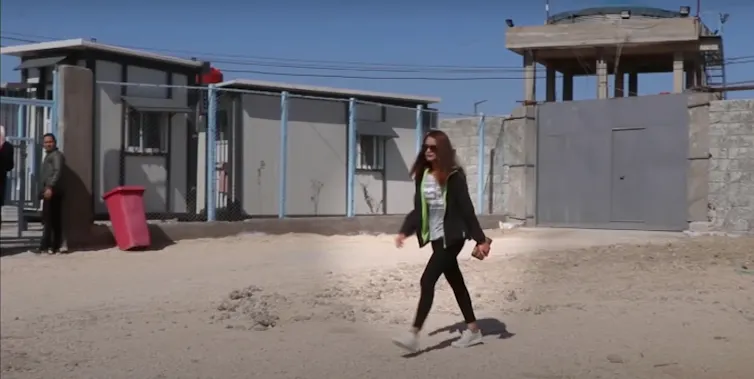 Shamima Begum walking across sand with detention camp buildings in the background