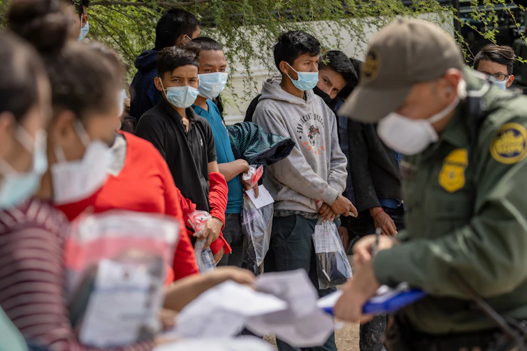 Customs agent in olive green with badges questions a group of women, while teenage boys in masks look on