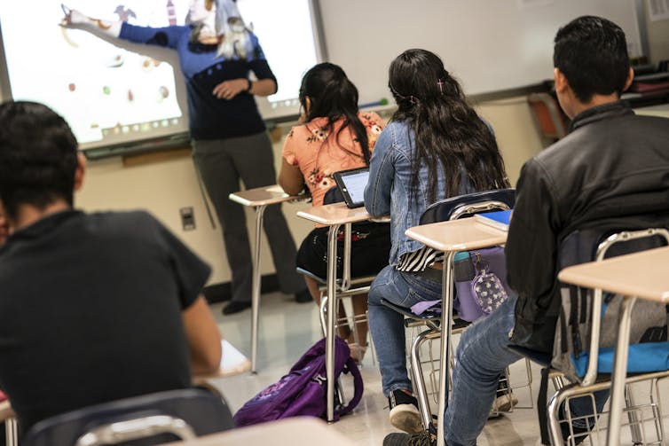 Los niños se sientan en un aula con un maestro apuntando a una pizarra en la parte delantera