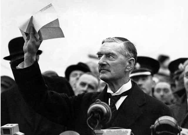 Elderly man in 1930s in the middle of a crowd of people waving a piece of paper in his right hand. 