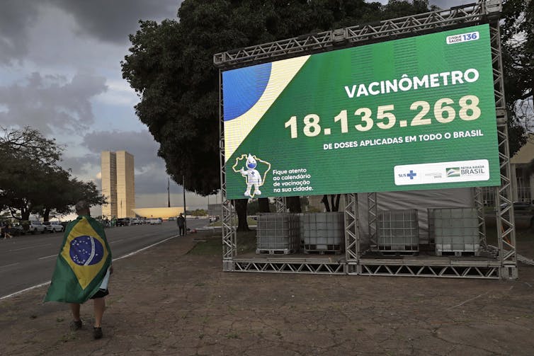 La cape du drapeau de l'homme au Brésil passe devant un panneau d'affichage montrant un total cumulatif du déploiement du vaccin au Brésil.