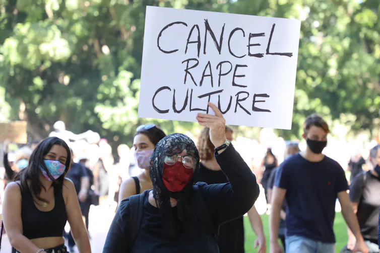 Masked protestor in black hoodie holds up card at protest that says 'CANCEL RAPE CULTURE'