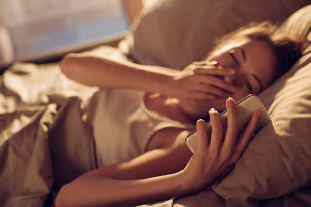 Woman lying in bed yawning and looking at phone