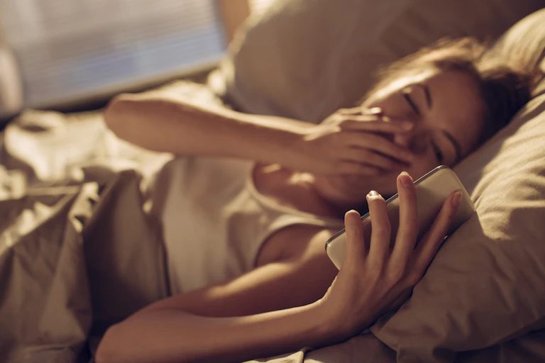 Woman lying in bed yawning and looking at phone
