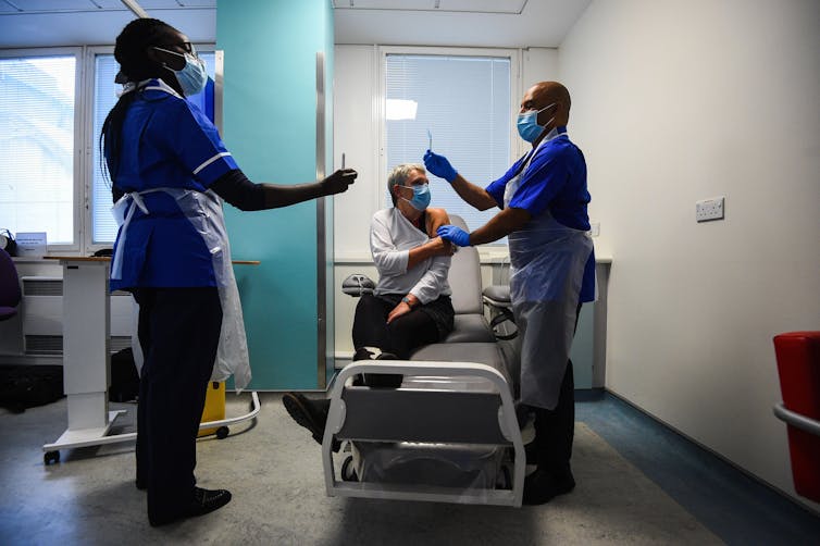 Two health workers administer a vaccine to a woman seated on a hospital bed as part of the Novavax vaccine trial.