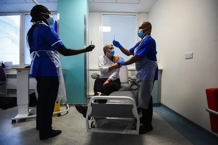 Two health workers administer a vaccine to a woman seated on a hospital bed as part of the Novavax vaccine trial.