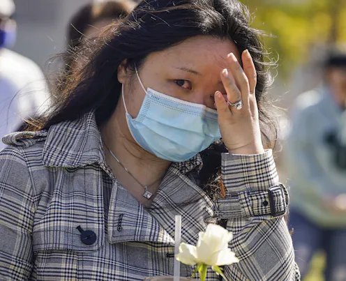 An Asian woman in a face mask with a white flower wiping a tear from her eye